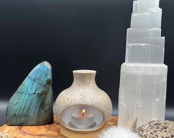 Puede incluir: Una piedra de labradorita pulida con iridiscencia azul, un portavelas de cerámica moteada con una vela encendida y una torre de selenita blanca de varios niveles. Estos objetos están dispuestos sobre una tabla de madera con madera de cholla.
