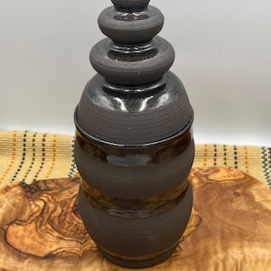 May include: A dark charcoal-colored ceramic jar with a matching lid. The jar features a cylindrical body with horizontal ridges and a smooth, glossy finish. The lid is tiered with three distinct, rounded sections, culminating in a pointed top.