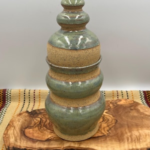 May include: A ceramic jar with a lid, featuring a stacked, bulbous silhouette. The piece is finished with a speckled, sandy-colored clay base and a glossy, sage-green glaze applied to the rounded sections and the pointed finial top.
