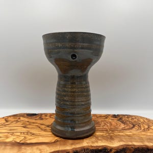 May include: A dark brown ceramic hookah bowl with a wide, flared top and a narrow, cylindrical base. The exterior features horizontal ridges and a smooth, glossy glaze with subtle black and bronze undertones. A single circular hole is visible.