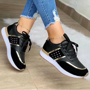 May include: Black and gold sneakers with white soles. The sneakers have black laces and a gold accent stripe. The toe and heel are black suede. The sneakers are worn with blue jeans.