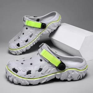May include: A pair of gray clogs with a textured design and neon green accents. The clogs have a strap with a black buckle and the word "abba" printed on the strap. The soles have a rugged, textured design.