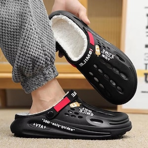 May include: Black clogs with a white, fuzzy interior and a red strap. The clogs have a perforated design and feature the word "SMILE" and other text. The sole is thick and the clogs appear to be made of a rubber-like material.