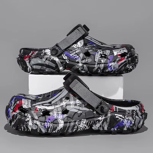 May include: A pair of black, grey, and white clogs with a graffiti-style design. The clogs have adjustable grey straps and are made of a durable material. The design includes splashes of red and purple.