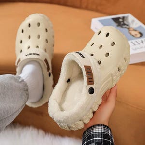 May include: Cream-colored clogs with a fuzzy interior and a strap that reads "MON". The clogs have multiple ventilation holes and a brown accent on the strap. The sole is textured for grip. The clogs are designed for comfort and warmth.