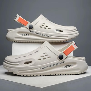 May include: Two pairs of off-white clogs with a sporty design. Each clog features a strap with orange accents, multiple ventilation holes, and the text "+VTA+" and "код-г-1885". The clogs have a thick, textured sole.