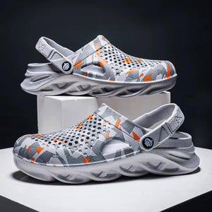 May include: A pair of grey and orange patterned clogs. The clogs feature a perforated upper design and a strap that can be worn around the heel or folded forward. The soles are white with a wavy design.