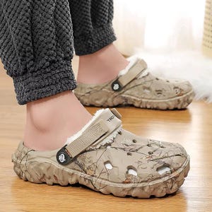 May include: Tan and brown camouflage-patterned clogs with a faux fur lining. The clogs have a strap that can be worn around the heel or across the top of the foot. The soles are thick and textured.