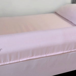 May include: A pink bed with a white bed skirt and a pink bed bumper. The bed bumper has a white zipper and the text "Slumber Bumper by Ziggysty Zag" printed on it.