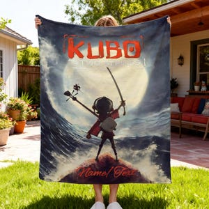 Puede incluir: Una manta rectangular de forro polar con el p&oacute;ster de la pel&iacute;cula Kubo y las dos cuerdas. El dise&ntilde;o muestra a un personaje con una espada y un shamisen frente a una gran luna y olas del mar. El texto dice Kubo and the Two Strings y Name/Text.