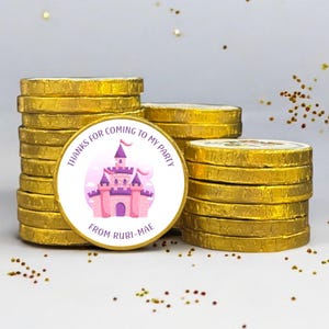 May include: Gold-wrapped chocolate coins stacked, with a circular sticker showing a pink castle and the text "THANKS FOR COMING TO MY PARTY FROM RUBI-MAE". The coins are arranged in stacks, with some glitter scattered around.