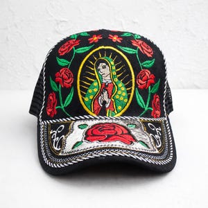 May include: Black trucker hat with embroidered design. Features a central oval image of a religious figure, surrounded by red roses and green leaves. The brim has a decorative floral design.