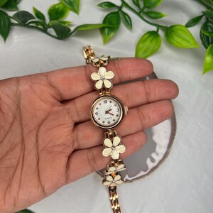 Puede incluir: Un reloj de cuarzo dorado con esfera blanca y detalles florales. La correa del reloj presenta una serie de eslabones con forma de flor blanca. La esfera del reloj tiene peque&ntilde;os puntos que marcan las horas.