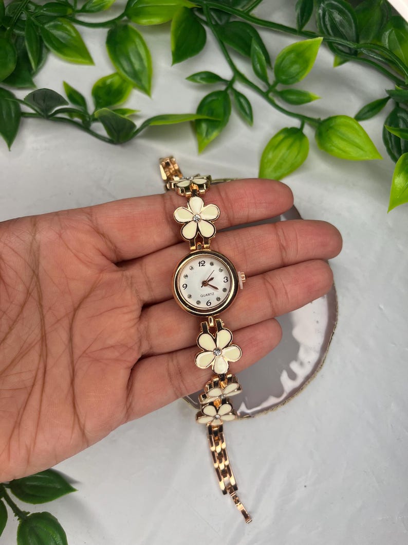 Puede incluir: Un reloj de cuarzo dorado con esfera blanca y detalles florales. La correa del reloj presenta una serie de eslabones con forma de flor blanca. La esfera del reloj tiene peque&ntilde;os puntos que marcan las horas.