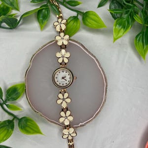 Puede incluir: Un reloj de cuarzo dorado con esfera blanca y dise&ntilde;o floral. La correa del reloj presenta detalles de flores color crema con centros de cristal. El reloj se exhibe sobre una superficie ovalada gris claro.