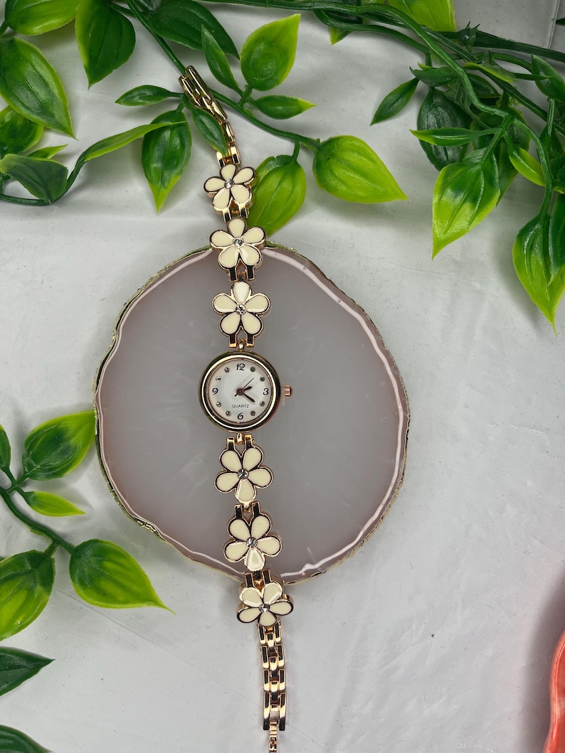 Puede incluir: Un reloj de cuarzo dorado con esfera blanca y dise&ntilde;o floral. La correa del reloj presenta detalles de flores color crema con centros de cristal. El reloj se exhibe sobre una superficie ovalada gris claro.