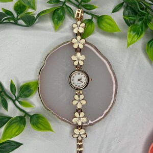 Puede incluir: Un reloj de pulsera dorado con esfera blanca y detalles florales. La correa del reloj presenta una serie de flores color crema con centros dorados. La esfera muestra los n&uacute;meros del 1 al 12. El reloj mide aproximadamente 18 cm de largo.
