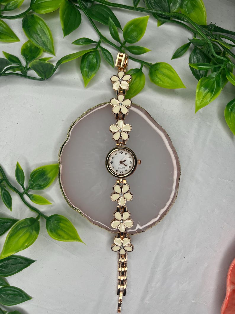 Puede incluir: Un reloj de pulsera dorado con esfera blanca y detalles florales. La correa del reloj presenta una serie de flores color crema con centros dorados. La esfera muestra los n&uacute;meros del 1 al 12. El reloj mide aproximadamente 18 cm de largo.