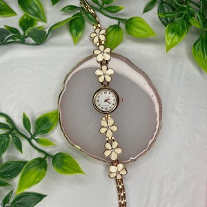 Puede incluir: Un reloj de pulsera dorado con esfera blanca y dise&ntilde;o floral. La correa del reloj presenta detalles de flores color crema. El reloj se exhibe sobre una superficie ovalada de color claro con follaje verde en el fondo.