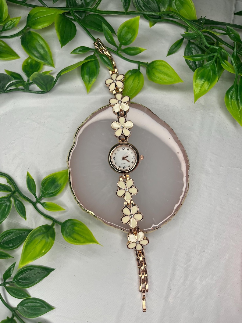 Puede incluir: Un reloj de pulsera dorado con esfera blanca y dise&ntilde;o floral. La correa del reloj presenta detalles de flores color crema. El reloj se exhibe sobre una superficie ovalada de color claro con follaje verde en el fondo.