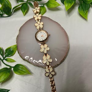 Puede incluir: Un reloj en tono oro rosa con esfera blanca y detalles florales. La correa del reloj presenta eslabones en forma de flor de color crema. El reloj se exhibe sobre una piedra decorativa rosa claro y blanca, con hojas verdes en el fondo.