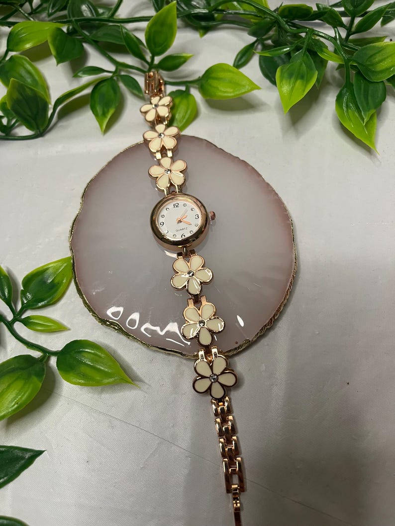 Puede incluir: Un reloj en tono oro rosa con esfera blanca y detalles florales. La correa del reloj presenta eslabones en forma de flor de color crema. El reloj se exhibe sobre una piedra decorativa rosa claro y blanca, con hojas verdes en el fondo.