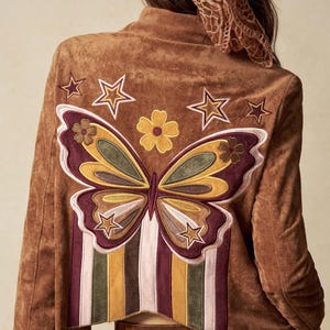 Puede incluir: Chaqueta de ante marrón con un gran bordado de mariposa y estrellas en la espalda. El diseño de la mariposa presenta detalles en burdeos, amarillo y verde. La chaqueta tiene un cuello alto y rayas verticales.