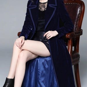 May include: A long, navy blue velvet coat with a contrasting trim. The coat is worn over a black top and shorts. The model is seated in a wooden chair, wearing black ankle boots. The coat's lining is a lighter shade of blue.