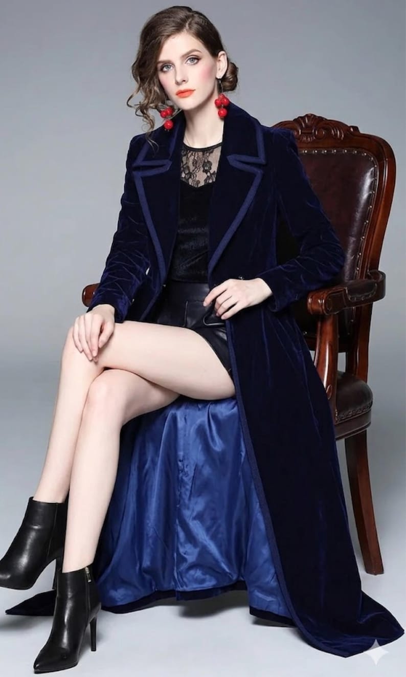 May include: A long, navy blue velvet coat with a contrasting trim. The coat is worn over a black top and shorts. The model is seated in a wooden chair, wearing black ankle boots. The coat's lining is a lighter shade of blue.