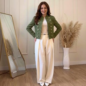 Puede incluir: Una chaqueta verde oliva de estilo militar con botones dorados se combina con pantalones de pierna ancha color crema. La chaqueta tiene un cuello alto y varias filas de botones. Una camiseta blanca y un cinturón delgado completan el atuendo. Un espejo de cuerpo entero y un jarrón con hierba alta están en el fondo.