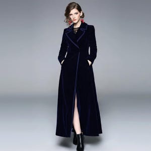 May include: A long, navy blue velvet coat with a notched collar and a front slit. The coat has long sleeves and pockets. The model is wearing black boots. The coat is a stylish outerwear garment.