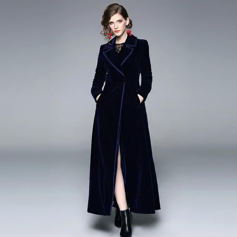 May include: A long, navy blue velvet coat with a notched collar and a front slit. The coat has long sleeves and pockets. The model is wearing black boots. The coat is a stylish outerwear garment.