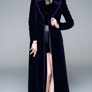 May include: A long, navy blue velvet coat with a contrasting dark blue trim. The coat has a classic lapel collar and a floor-length design. The model is wearing a black top and black boots. The coat is a stylish outerwear piece.