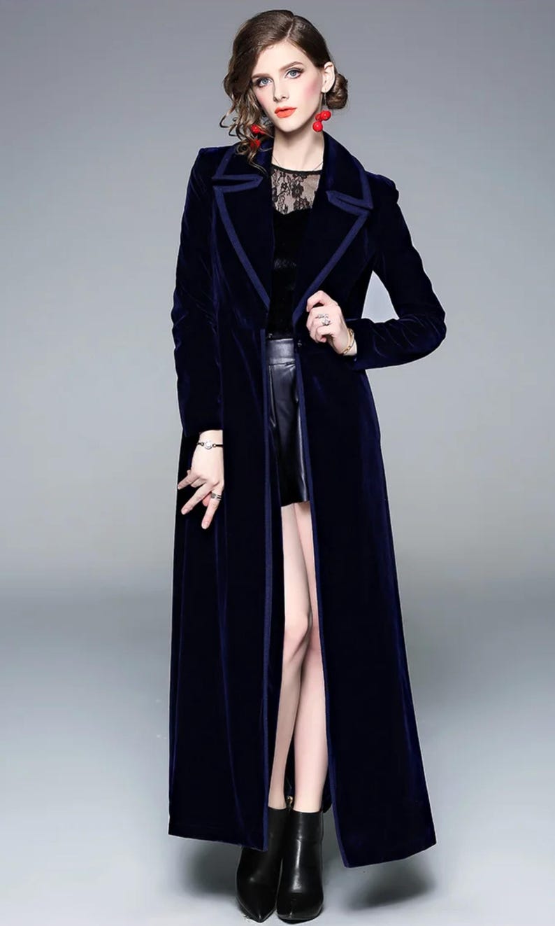 May include: A long, navy blue velvet coat with a contrasting dark blue trim. The coat has a classic lapel collar and a floor-length design. The model is wearing a black top and black boots. The coat is a stylish outerwear piece.