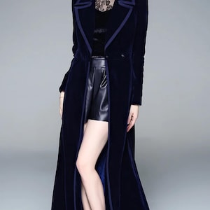 May include: A long, navy blue velvet coat with a contrasting dark blue trim. The coat is floor-length and features a notched collar and a single-button closure. The model is wearing black leather shorts and black ankle boots. The coat's design is elegant and sophisticated.