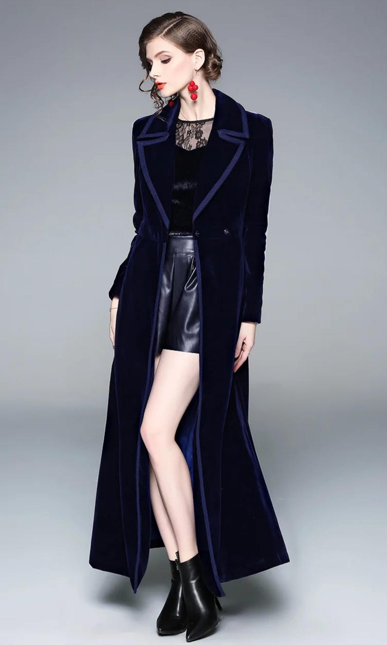 May include: A long, navy blue velvet coat with a contrasting dark blue trim. The coat is floor-length and features a notched collar and a single-button closure. The model is wearing black leather shorts and black ankle boots. The coat's design is elegant and sophisticated.