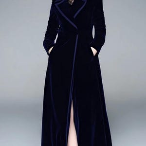 May include: A long, navy blue velvet coat with a tailored design. The coat features a notched collar, long sleeves, and a high slit in the front. The coat has pockets and is worn over a black top.