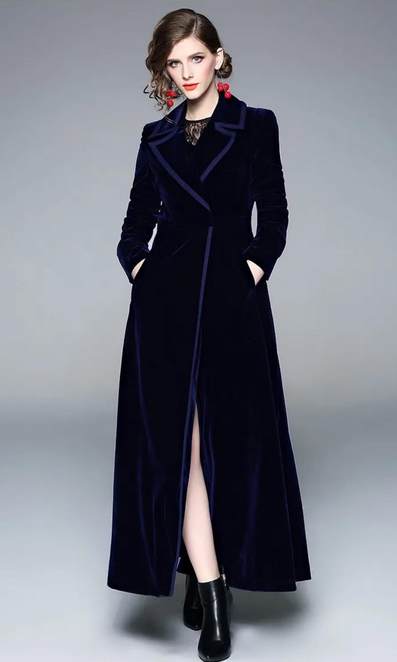 May include: A long, navy blue velvet coat with a tailored design. The coat features a notched collar, long sleeves, and a high slit in the front. The coat has pockets and is worn over a black top.