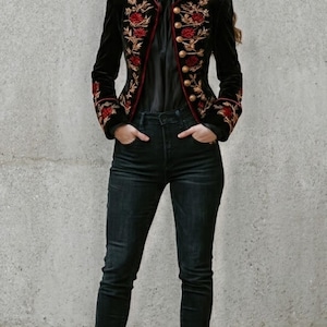 May include: A black velvet jacket featuring intricate red floral embroidery and gold-toned buttons, worn with a black blouse, dark denim jeans, and black leather ankle boots. The jacket has a high collar and structured shoulders.