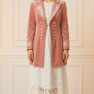 May include: A dusty rose velvet coat with ornate embroidery on the collar and cuffs. The coat features a row of buttons down the front and is worn over a white lace dress. The outfit is completed with gold-colored sandals.