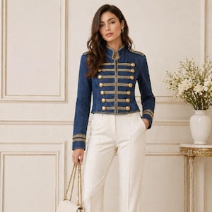 Könnte beinhalten: Dunkelblaue Jeansjacke mit goldenen Details, darunter Knöpfe und Verzierungen. Die Jacke hat einen militärischen Stil mit hohem Kragen und goldenen Streifen an den Ärmeln. Sie wird mit einer weißen Hose und einer weißen Handtasche getragen.