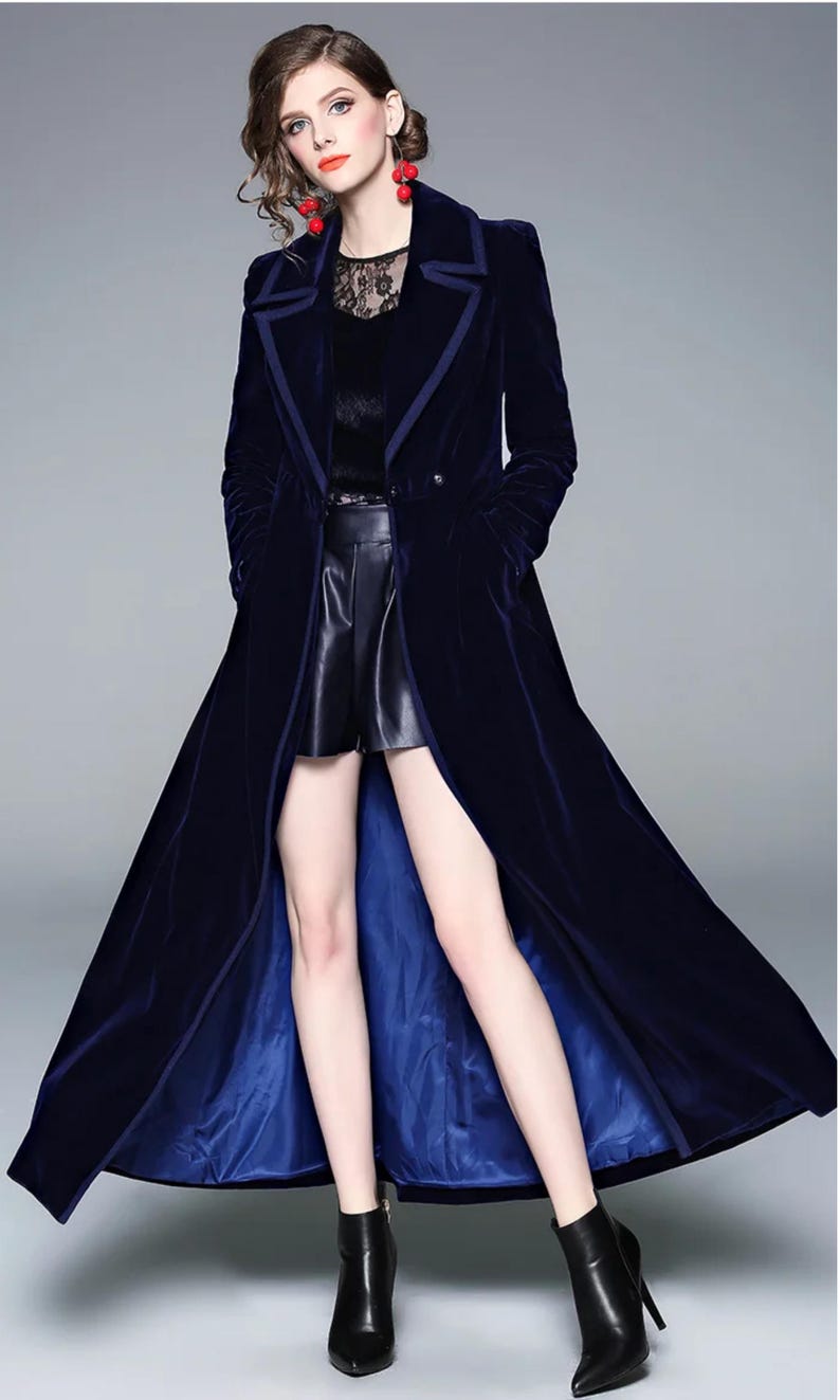 May include: A long, navy blue velvet coat with a contrasting blue lining. The coat features a classic lapel and a double-breasted design. The model is wearing black leather shorts and black ankle boots.