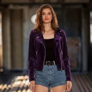 May include: A deep purple velvet jacket with a zipper closure and a black belt. The jacket is paired with denim shorts and a black top. The model is standing in a blurred background.