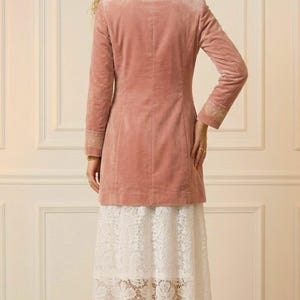 May include: A dusty rose velvet blazer with long sleeves and gold embroidered cuffs. The blazer is paired with a white lace skirt. Gold high heels complete the outfit, showcasing a sophisticated, feminine style.