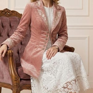 May include: A dusty rose velvet blazer with ornate embroidery on the lapels and cuffs. The blazer features a row of decorative buttons and is paired with a white lace dress. The image showcases a vintage-style chair and high-heeled sandals.