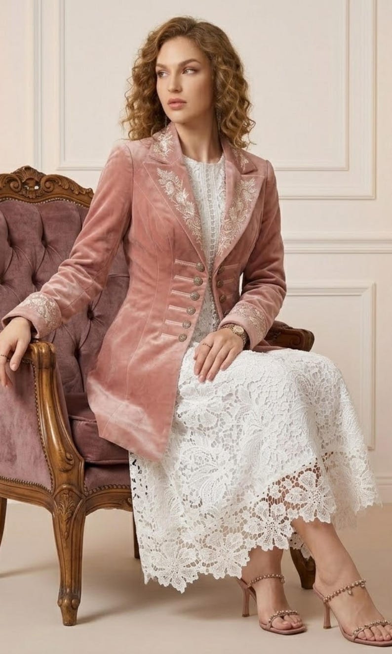 May include: A dusty rose velvet blazer with ornate embroidery on the lapels and cuffs. The blazer features a row of decorative buttons and is paired with a white lace dress. The image showcases a vintage-style chair and high-heeled sandals.