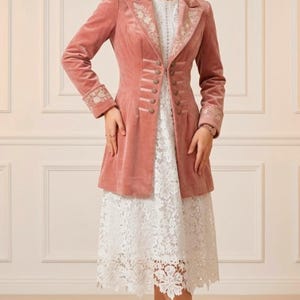 May include: A dusty rose velvet coat with ornate embroidery on the collar and cuffs. The coat features a row of decorative buttons and is worn over a white lace dress. The outfit is completed with strappy sandals.