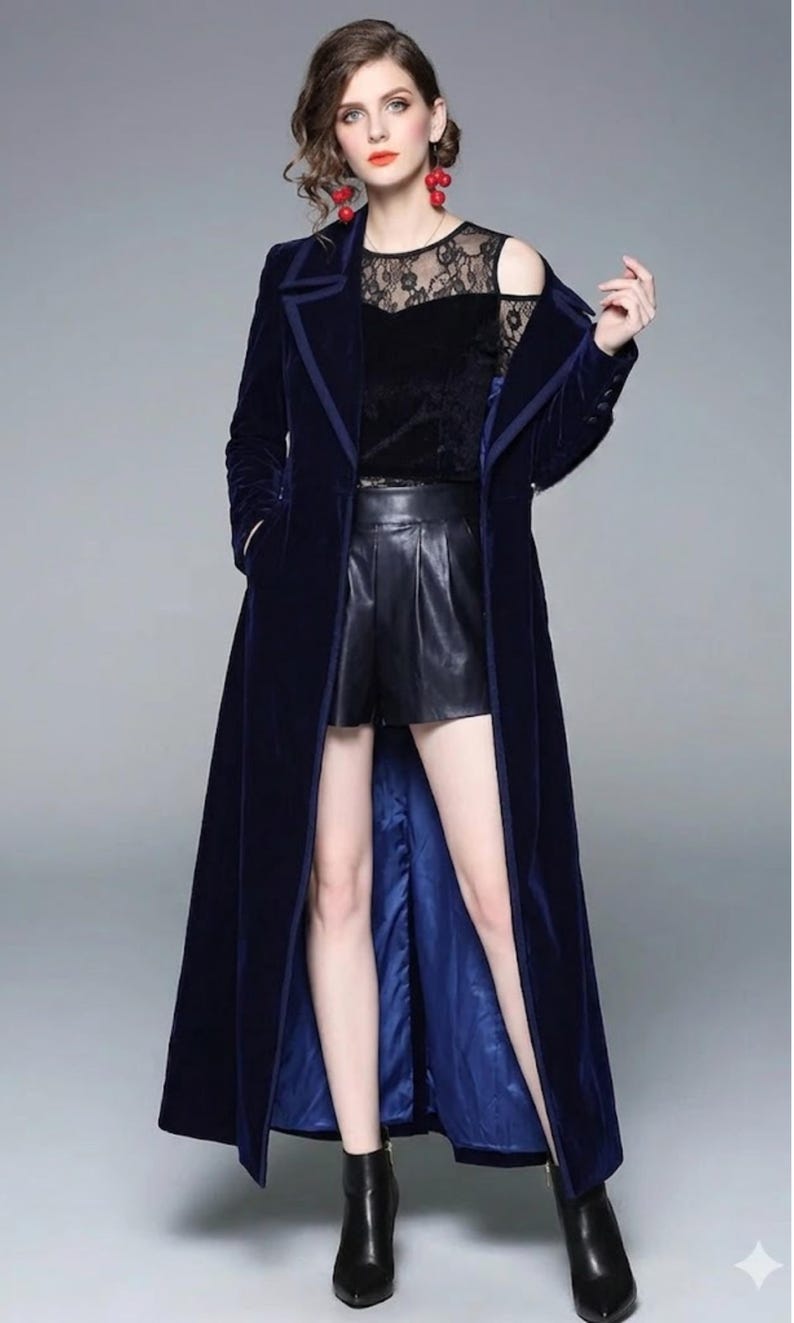 May include: A long, navy blue velvet coat with a contrasting blue lining and trim. The coat is worn over a black lace top and black shorts. The model is also wearing black ankle boots. The coat has a classic, elegant design.