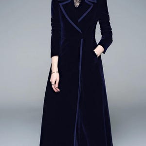 May include: A long, navy blue velvet coat with a contrasting dark blue trim. The coat features a notched collar, long sleeves, and a front closure. It has a tailored fit and a floor-length hemline, creating an elegant silhouette.