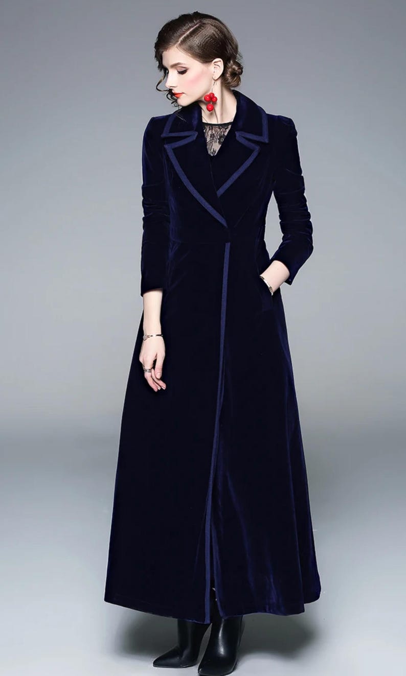 May include: A long, navy blue velvet coat with a contrasting dark blue trim. The coat features a notched collar, long sleeves, and a front closure. It has a tailored fit and a floor-length hemline, creating an elegant silhouette.